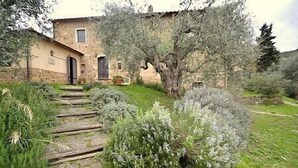 Un bâtiment en pierre à l’aspect rustique, avec un escalier en bois menant à l’entrée et un jardin bien entretenu agrémenté de divers arbustes et d’un grand arbre.