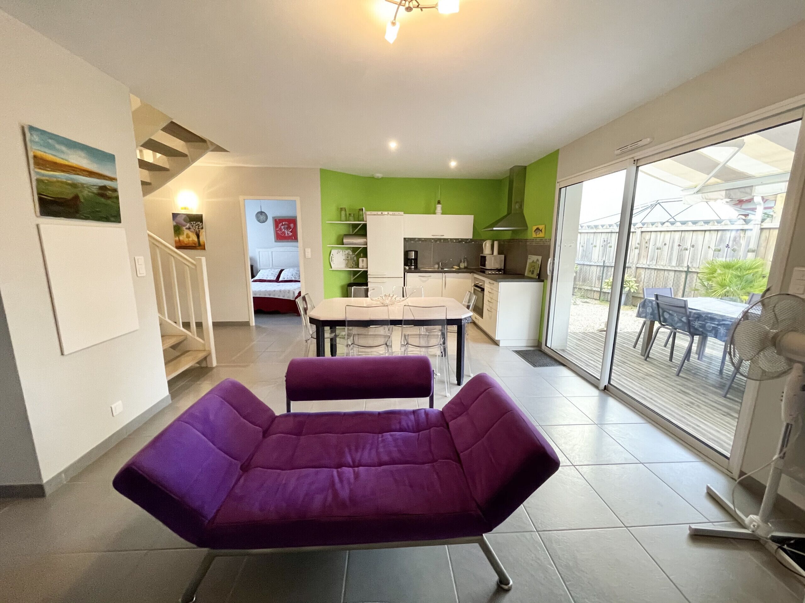 A modern living room with a purple sofa, a dining area, a kitchen, and a sliding glass door leading to a patio.