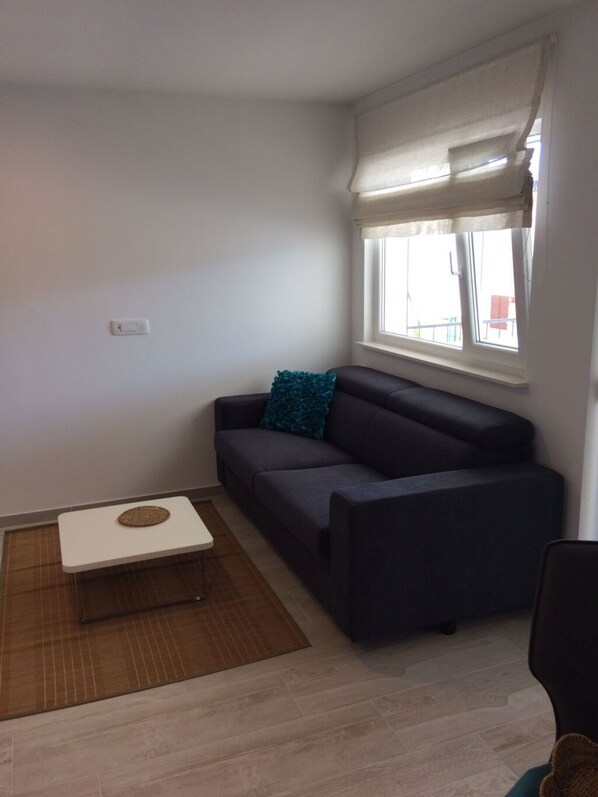 A small, modern living room with a dark grey sofa, a white coffee table, and a window with white curtains.