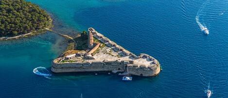 An aerial view of a fortified island with a lighthouse, surrounded by clear blue waters and boats.