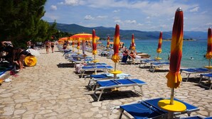 Une plage bordée de rangées de chaises longues et de parasols, une promenade dallée et une vue sur la mer et les montagnes en arrière-plan.