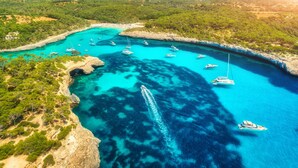 An aerial view of a coastal area with clear turquoise waters, boats, and rocky terrain.