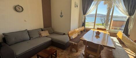 A living room with a sofa, wooden table, and chairs, a clock on the wall, and a view of the sea through large windows.
