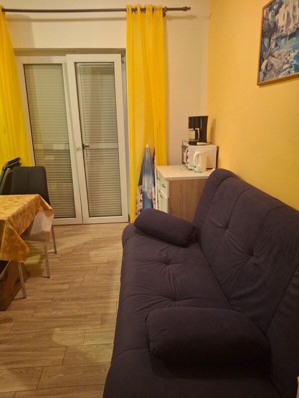 Una sala de estar con un sofá oscuro, una mesita con mantel amarillo, una silla y una zona de cocina con un microondas.