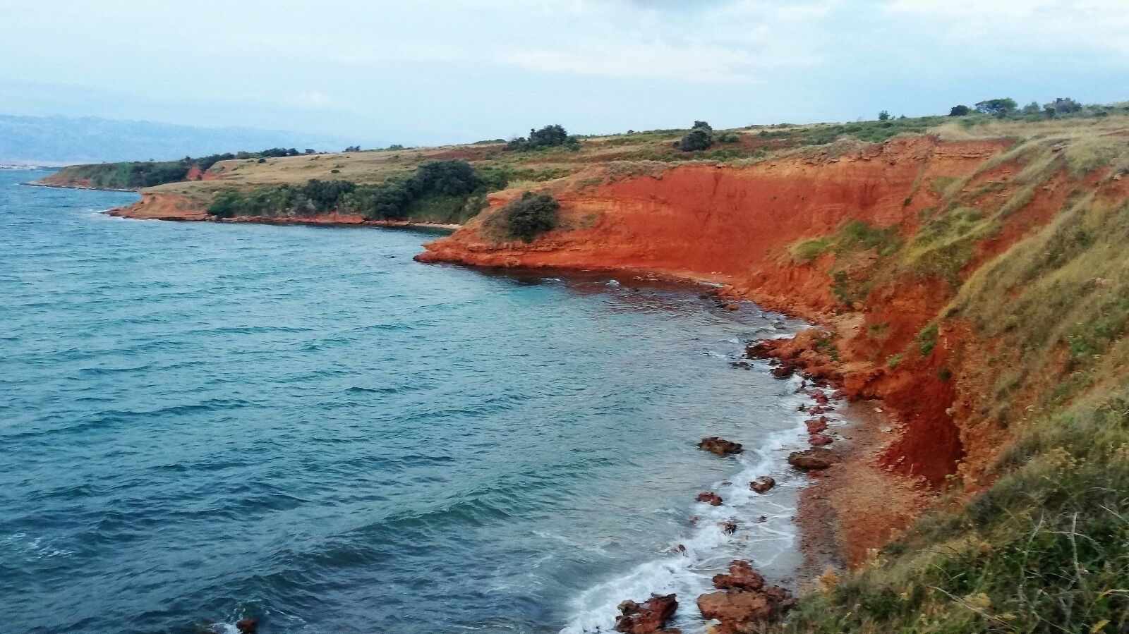 A coastal landscape with a red cliff and a clear blue sea.