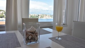 A table with a glass vase of seashells and a glass of orange juice, a view of a coastal town, and a mountain range in the background. - Holiday Home in Sumartin with Seaview, Air condition, WIFI, Washing machine (682-2) (Sumartin)