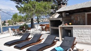 A poolside area with lounge chairs, a stone building with a chimney, and a view of the sea and buildings in the background. - Holiday Home in Sumartin with Seaview, Air condition, WIFI, Washing machine (682-2) (Sumartin)