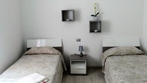 Camera da letto con due letti, un comodino con lampada e due mensole a parete con oggetti decorativi.