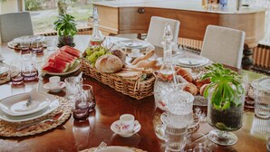 Una mesa de comedor dispuesta con platos, vasos y diversos alimentos, rodeada de sillas y con vista a zonas verdes.