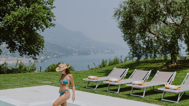 Una mujer en traje de baño al lado de una piscina con sillones de descanso y vistas a montañas.