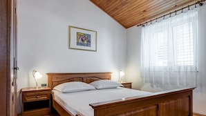 A bedroom with a wooden bed, two bedside tables with lamps, a framed picture on the wall, and a window with sheer curtains.