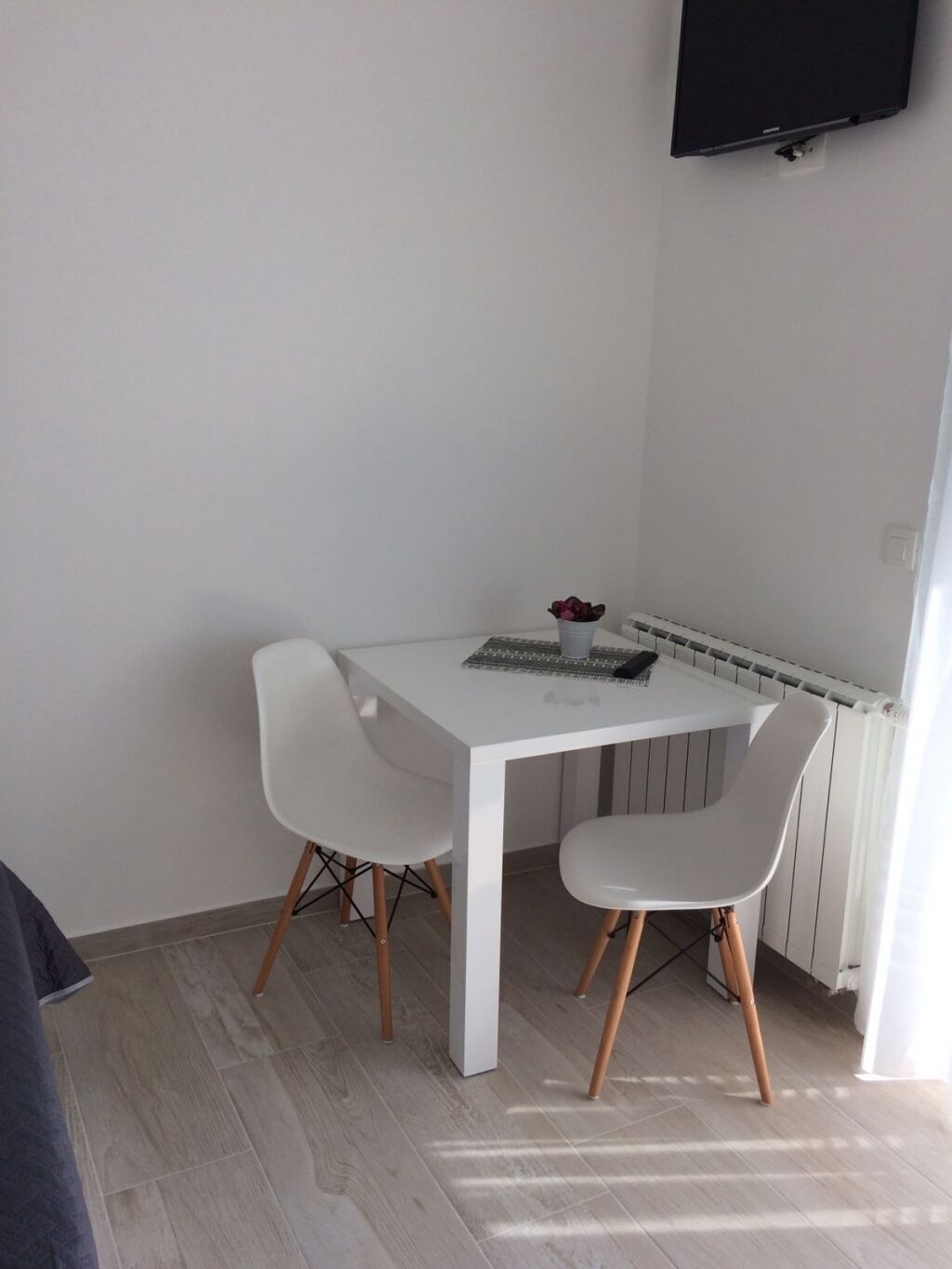 A small, white dining area with a table and two chairs, a radiator below, and a TV mounted on the wall.