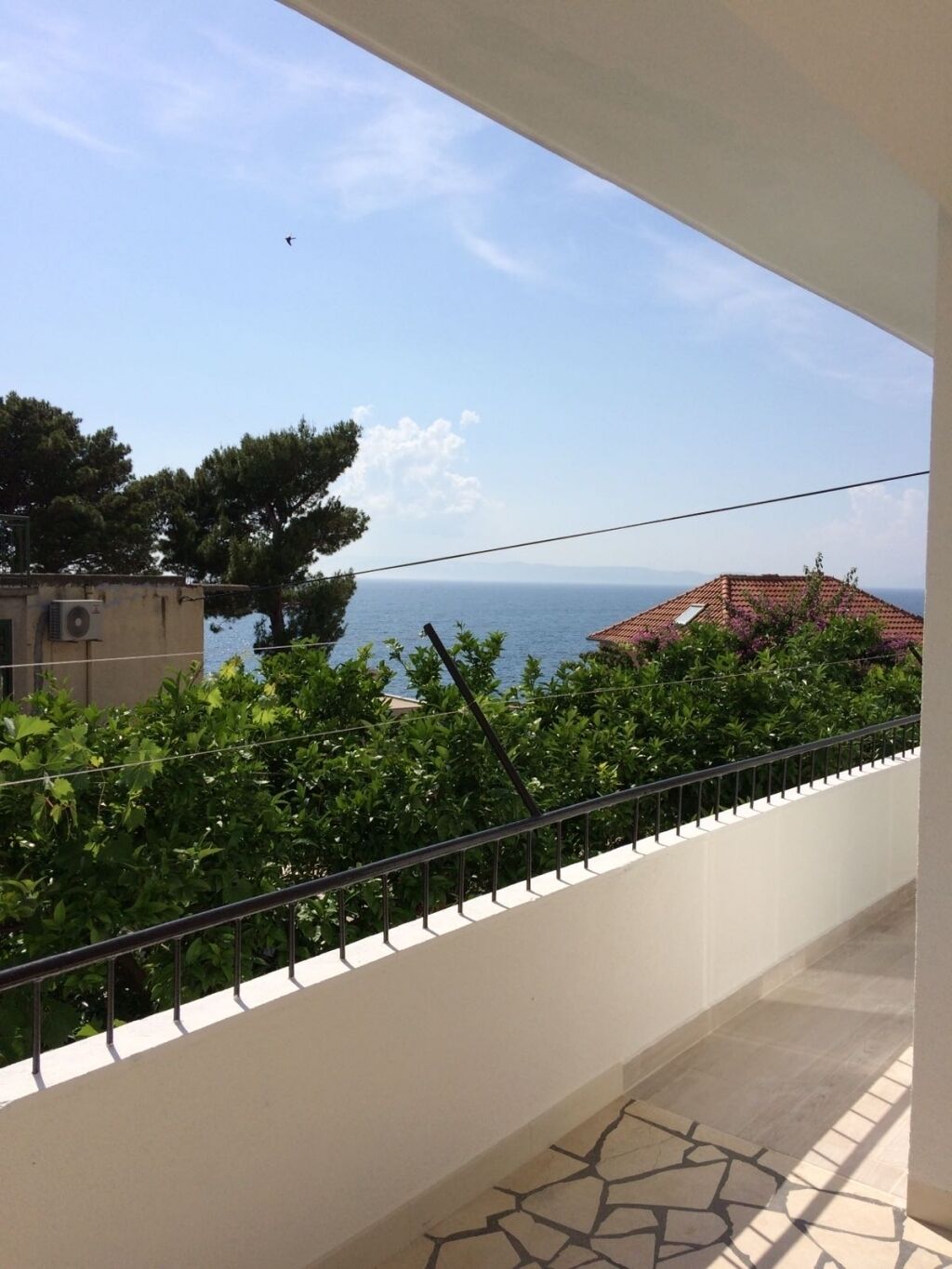 Ein Balkon mit Blick auf das Meer, Grünflächen und ein Gebäude mit rotem Dach.