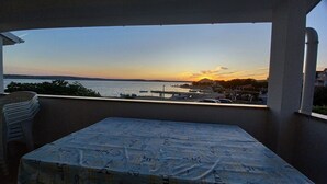 Un balcon avec une table et des chaises, donnant sur une marina où sont amarrés des bateaux, et offrant une vue sur le coucher de soleil.