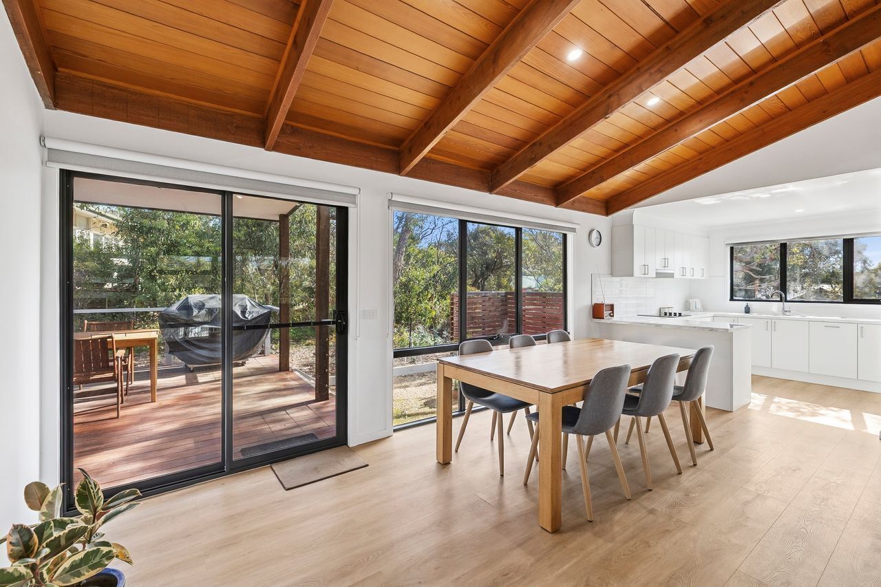 Une cuisine moderne avec des armoires blanches et un grand îlot central, un coin repas avec une table et des chaises en bois, et une porte-fenêtre coulissante donnant sur un patio extérieur équipé d’un barbecue.