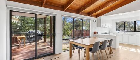 A modern kitchen with white cabinets and a large island, a dining area with a wooden table and chairs, and a sliding glass door leading to an outdoor patio with a barbecue grill.