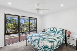 A bedroom with a large window, a bed with a leaf-patterned bedspread, a ceiling fan, and a nightstand with a lamp. - Nod Off On Noble (Anglesea)