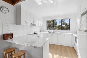 A modern kitchen with white cabinetry, a marble countertop, and a window providing a view of trees. - Nod Off On Noble (Anglesea)