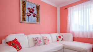 Un divano bianco componibile con cuscini floreali, una parete rosa, un quadro appeso sopra e una finestra con tende leggere.