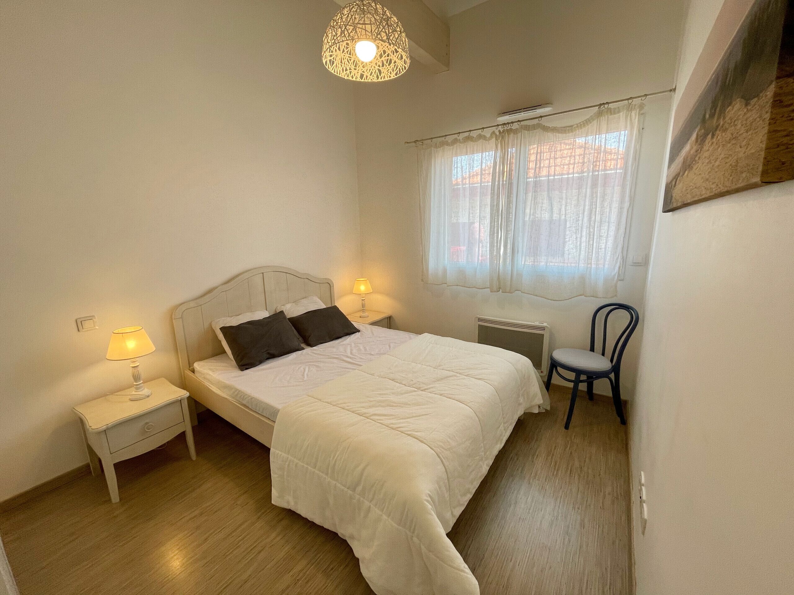 A bedroom with a bed, two bedside lamps, a chair, a radiator, and a window with curtains.