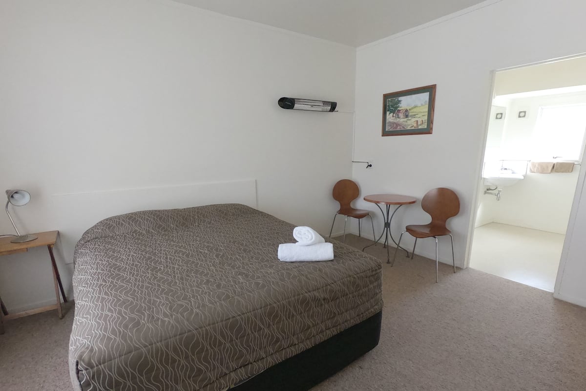 A bedroom with a large bed, a desk with a lamp, two chairs, a small table, and a painting on the wall.
