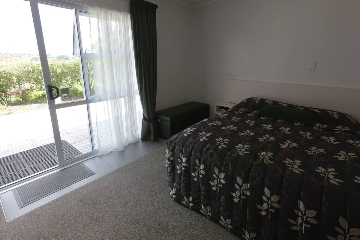 A bedroom with a bed, a small bedside table, a window with curtains, and a view of greenery outside.