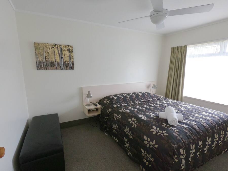 A hotel room with a bed, a nightstand, a lamp, a ceiling fan, a painting on the wall, and a window with curtains.