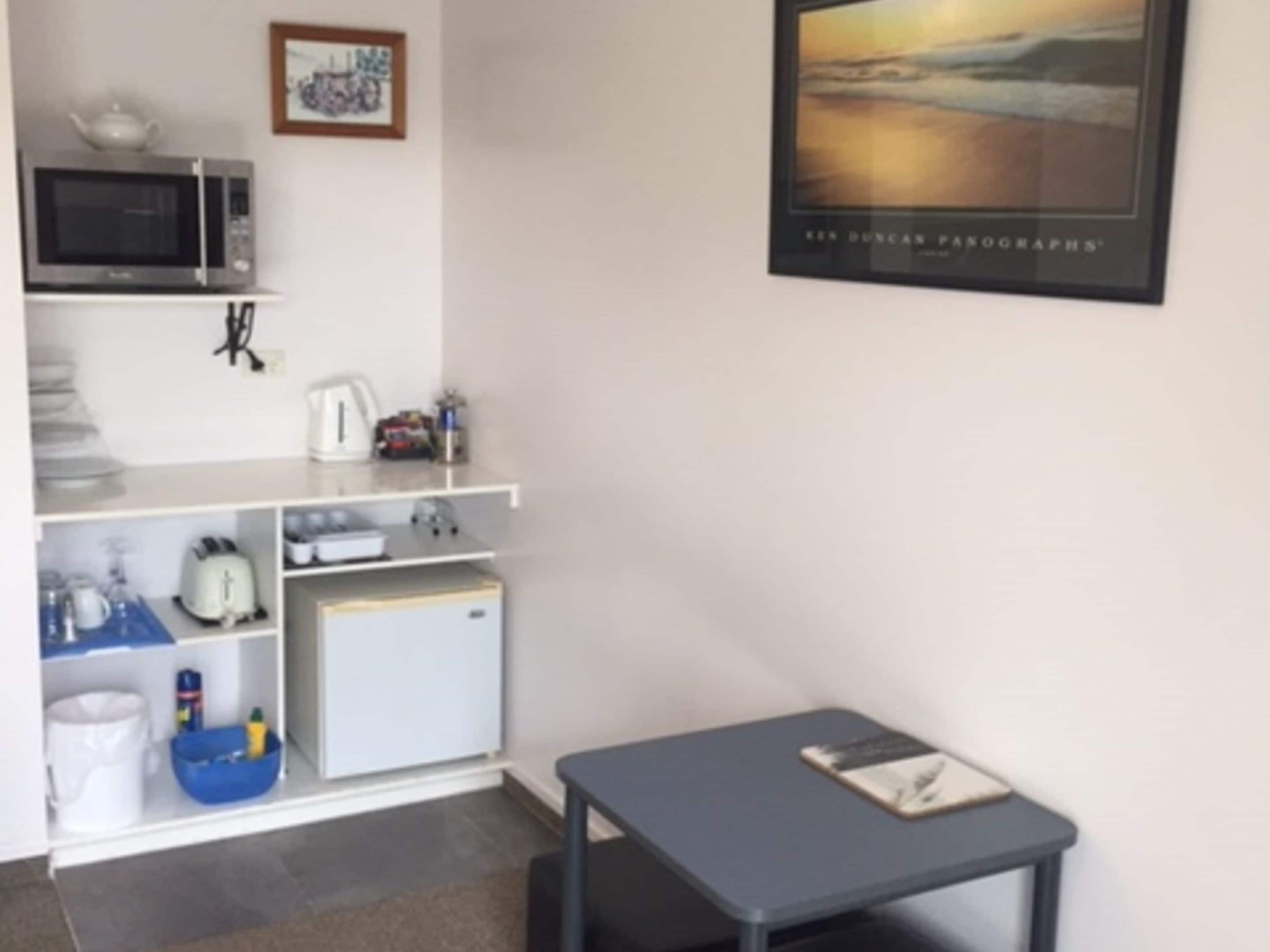 Een kleine kitchenette met een magnetron, waterkoker, broodrooster en koelkast. Er hangt een lijst met een foto aan de muur en er staat een klein tafeltje met een boek.
