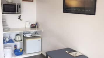 Een kleine kitchenette met een magnetron, waterkoker, broodrooster en koelkast. Er hangt een lijst met een foto aan de muur en er staat een klein tafeltje met een boek.