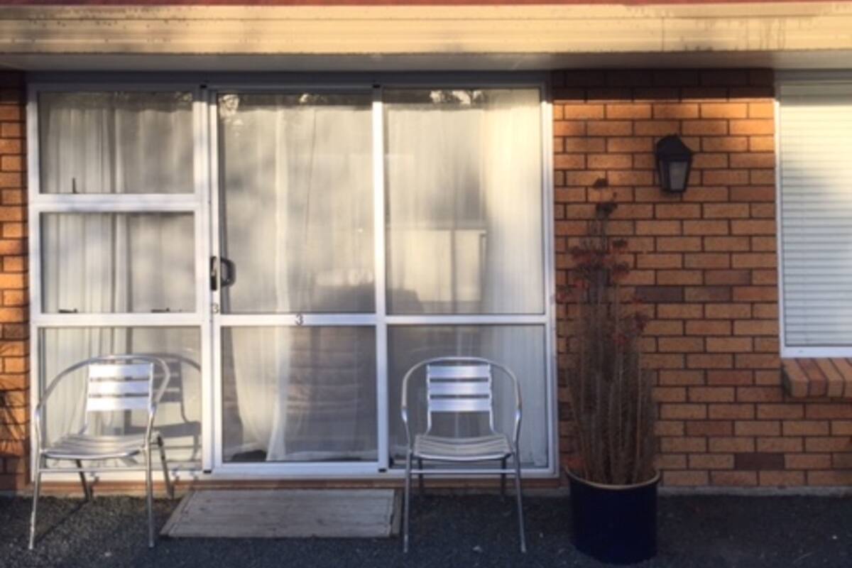 A building with a brick facade, a large sliding glass door with two metal chairs outside, and a potted plant to the right.