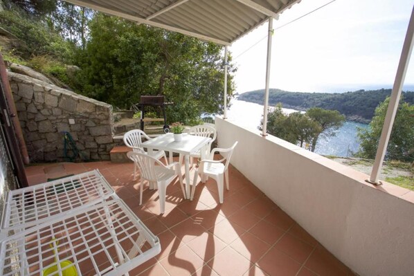 A covered patio with a table and chairs, overlooking a body of water and hills.