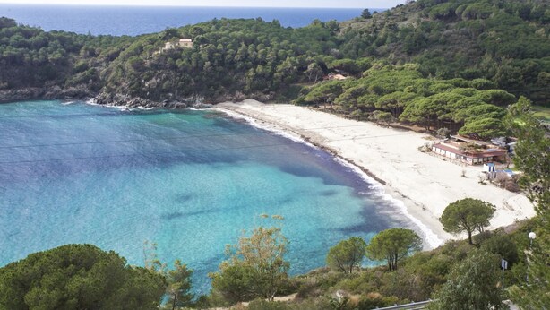 A coastal landscape with a beach, clear blue waters, and dense green hills.