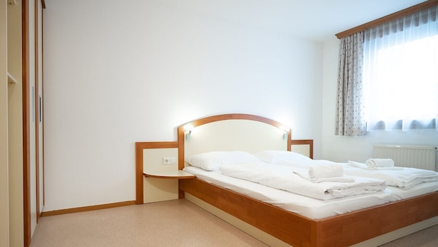 Een net opgemaakt bed met een houten hoofdbord en nachtkastje, een radiator en een raam met gordijnen.