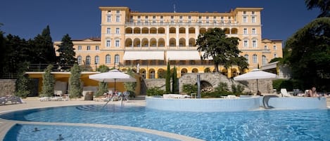 Un vaste espace piscine comprenant plusieurs piscines et des chaises longues, entouré d’un bâtiment doté de nombreux balcons et d’un pont en pierre.