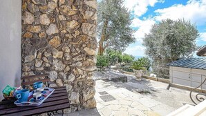 Un muro de piedra, asientos exteriores con una bandeja de vasos y comida, y vistas a árboles y un edificio al fondo.