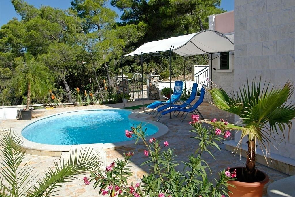 A swimming pool with blue water, surrounded by potted plants and flowers. There are two blue lounge chairs under a white umbrella.