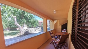 A balcony with a view of a tree and a stone wall, a wooden table with chairs, and a striped beach chair.