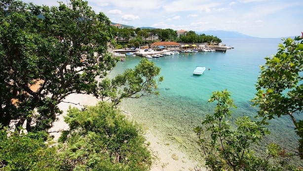 Vista costera con aguas transparentes, una playa de arena y un puerto deportivo con barcos.