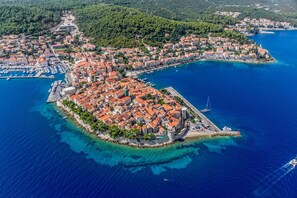 An aerial view of a coastal town with a prominent square surrounded by buildings with red-tiled roofs, a marina with boats, and a clear blue sea.