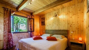 A wooden cabin bedroom with a bed, two pillows, a nightstand, a window with curtains, and a wall-mounted light.