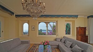 Ein Wohnzimmer mit einem großen Kronleuchter, zwei Sofas, einem Couchtisch mit Blumenvase und Blick durch Bogenfenster.