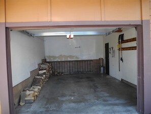 Un garage avec un sol en béton, une rambarde en métal et une pile de bois de chauffage appuyée contre le mur.