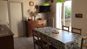 A dining area with a table covered in a tropical-themed tablecloth, wooden chairs, a wooden cabinet, a television on a shelf, and a clock on the wall. - House with 3 bedrooms, 8 people, Lacanau Océan (LACANAU OCEAN)