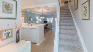 A modern kitchen with white cabinets, a central island, and a chandelier. A staircase leads to a living area with a blue wall and a dining table. - BELLA VITA ~ 2/1.5 located in the Truman Annex with a community pool! (Key West)