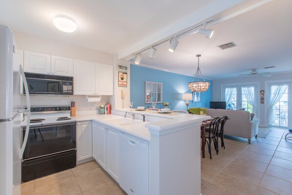 A modern kitchen with white cabinetry, a built-in oven, and a dishwasher. There is a dining area with a table and chairs, and a living room with a sofa and a chandelier. - BELLA VITA ~ 2/1.5 located in the Truman Annex with a community pool! (Key West)