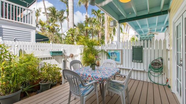 A covered outdoor patio with a table and chairs, surrounded by potted plants and a white picket fence. - BELLA VITA ~ 2/1.5 located in the Truman Annex with a community pool! (Key West)