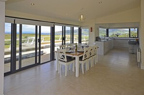 Un espace repas moderne avec une grande table dressée pour six personnes, de grandes fenêtres offrant une vue sur un terrain de golf et un coin cuisine donnant sur un espace verdoyant.