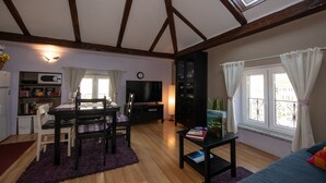 Ein gemütliches Wohnzimmer mit Essbereich, Holzbalken, einem Fernseher und einem Fenster mit Vorhängen.