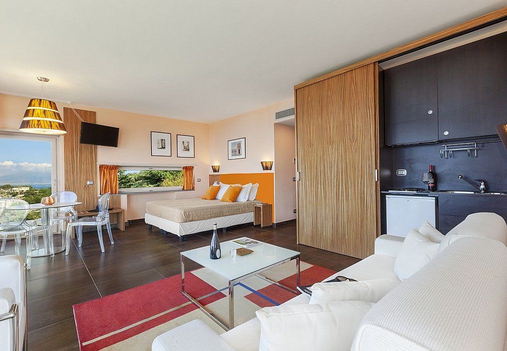 A modern hotel room with a bed, sofa, dining table, and kitchenette.
