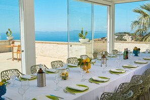 Une table dressée pour le dîner, avec vue sur la mer, des chaises d’extérieur et une plante en pot.